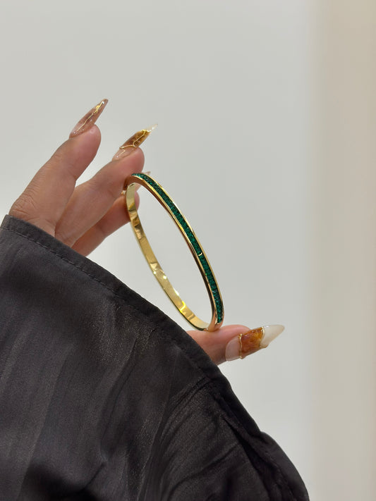 Gold bangle with green stones held by a hand against a neutral background