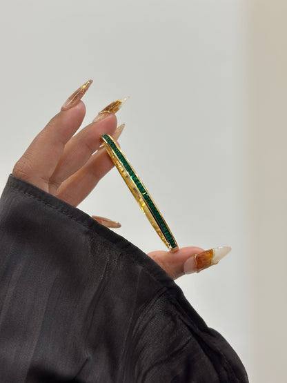 Gold and green bracelet held by a hand with long nails against a neutral background